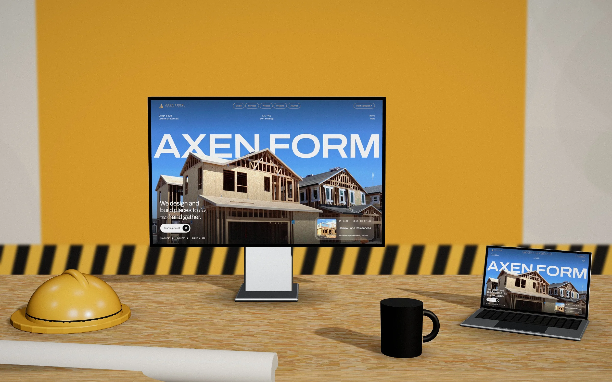The Axen Form website on a desktop monitor and a laptop, beside a yellow hard hat and a roll of plans on a timber desk